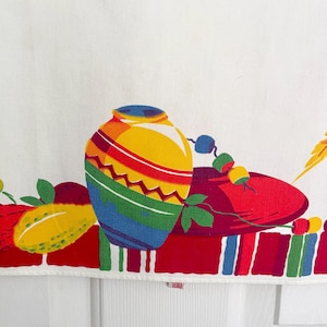 May include: A white fabric with a colorful, stylized design. The design features a large, multi-colored vase with a zig-zag pattern, a red sombrero, and other decorative elements. The border has vertical stripes in red, blue, and green.