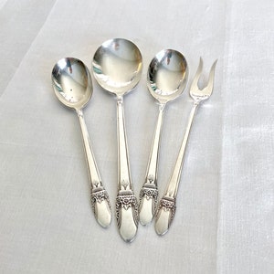 May include: A set of four silver-colored serving utensils: three spoons and one fork. The spoons have rounded bowls and long, slender handles. The fork has two tines. Each utensil features an ornate floral design at the base of the handle.