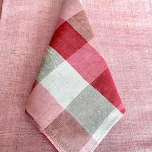 May include: A folded red, pink, and white checkered napkin. The napkin is made of a textured fabric and is placed on a matching pink surface. The checkered pattern is composed of squares in varying shades of red, pink, white, and gray.