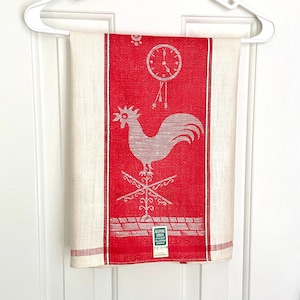 May include: A vintage kitchen towel with a red central panel featuring a rooster and clock design. The towel has a cream border with red stripes and is hanging on a white hanger. The towel is labelled "ALL PURE LINEN".