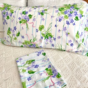 May include: A pair of white pillowcases with a blue and purple floral pattern. The pattern features small purple flowers with green leaves and pink ribbons.