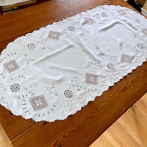 May include: White oval tablecloth with delicate embroidered floral and geometric designs. The tablecloth has a scalloped edge and is made of a lightweight fabric.