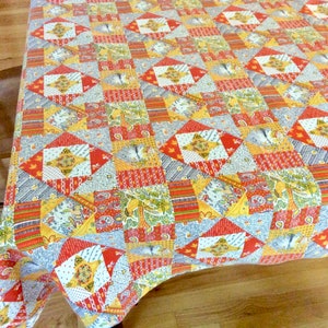 May include: A colorful patchwork quilt with a repeating pattern of diamonds and squares. The quilt features a mix of red, yellow, gray, and white fabrics with floral and geometric designs. The quilt is folded over a surface.