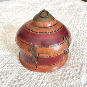 May include: A vintage, dome-shaped wooden box with a hinged lid and metal clasp. The box features a color scheme of reddish-brown, tan, and black, with decorative bands and a metal ring on top. The box is likely used for storage.