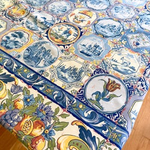 May include: A fabric tablecloth featuring a repeating tile pattern in shades of blue, yellow, and white. The design includes circular and octagonal motifs depicting landscapes, a hot air balloon, a tulip, and a camel, bordered by floral and fruit motifs.