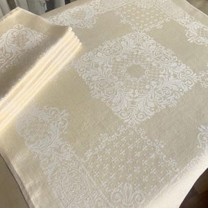 May include: A cream-colored tablecloth and matching napkins with a white, ornate floral pattern. The tablecloth is rectangular, and the napkins are stacked neatly to the side. The design is symmetrical and elegant.
