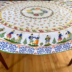 May include: A round white tablecloth featuring a colorful border of figures in traditional folk attire, including dancers and a bagpipe player. The center is decorated with a circular floral wreath pattern and scattered leaf motifs in orange, green, and blue.