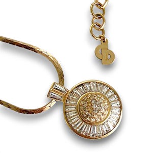 May include: Gold-toned necklace featuring a circular pendant with a central cluster of small, sparkling stones surrounded by larger, baguette-cut crystals. The necklace has a delicate chain and an adjustable clasp.