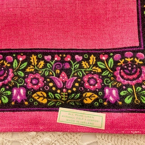 May include: A pink linen textile with a black border featuring a floral design in pink, purple, yellow, and green. The tag reads "All Pure Linen, Marshall Field & Company, Made in Ireland."