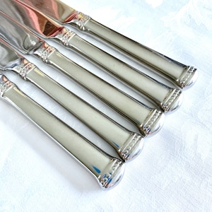 May include: A set of five stainless steel knives with a reflective, polished finish. The handles are a light, neutral color with a decorative beaded design near the blade and at the end of the handle. The knives are arranged diagonally on a white surface.