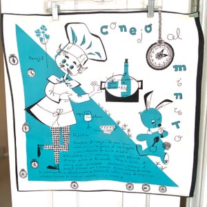 May include: A white and teal kitchen towel with a whimsical illustration of a chef and a rabbit preparing a dish. The towel features a recipe in Spanish, a clock, and the words "Conejo al Memento".