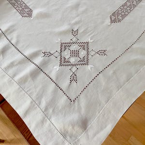 Peut inclure: Nappe en lin blanc cassé avec des motifs géométriques brodés complexes en marron foncé et blanc. Le design présente un motif carré central avec des éléments rayonnants et des bordures décoratives, créant un look classique et élégant.