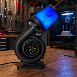 Puede incluir: Una lámpara negra con forma de turbocompresor con un filtro azul iluminado. La lámpara está sobre una superficie de madera, con un cable de alimentación negro. El fondo incluye herramientas y una impresora 3D, sugiriendo un taller.