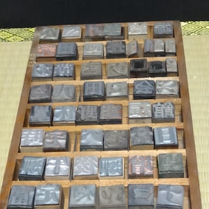 May include: A rectangular wooden typesetter tray containing numerous individual metal printing blocks. Each block features a unique raised Japanese kanji character on its top surface. The blocks are arranged in a grid pattern within the wooden dividers of the tray, showing a variety of metallic grey tones and textures.