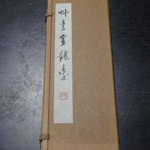 May include: A rectangular, tan-coloured book with a white label and black calligraphy. The book is bound with a light brown strap. A red seal is visible on the white label. The book appears to be of Asian origin.