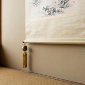Peut inclure: Une tenture murale suspendue présentant une peinture de paysage à l'encre monochrome sur papier crème. Le rouleau est lesté par une tige en bois foncé à la base, ornée de deux ornements sphériques décoratifs et de longs pompons jaune doré.
