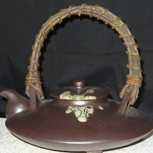 May include: A brown ceramic teapot with a woven handle. The teapot has a round body, a spout, and a lid with a small knob. The handle is made of light-coloured wood wrapped with darker twine. The lid has a light green floral design.
