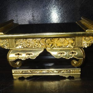 Può includere: Un supporto decorativo dorato e ornato, con una parte superiore e una base rettangolari. Il supporto presenta intagli floreali e volute decorative. Il supporto è probabilmente in metallo e ha una finitura lucida, alto circa 15 cm.