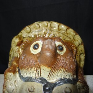 May include: A ceramic tanuki statue, a traditional Japanese raccoon dog figure. The statue features a detailed face with large eyes, a prominent nose, and a hat. The colors are primarily brown, tan, and cream, with some black accents. The figure has a textured surface.