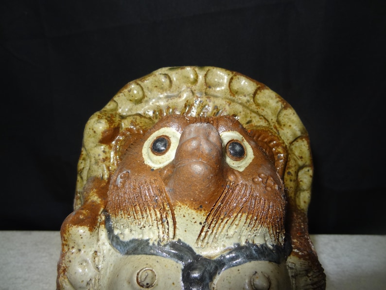 May include: A ceramic tanuki statue, a traditional Japanese raccoon dog figure. The statue features a detailed face with large eyes, a prominent nose, and a hat. The colors are primarily brown, tan, and cream, with some black accents. The figure has a textured surface.