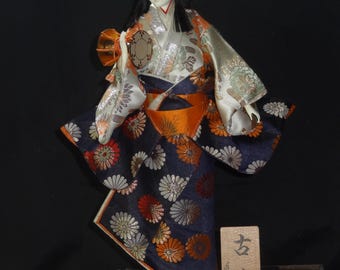 May include: A Japanese doll in a traditional kimono. The doll's kimono is navy blue with orange and white floral patterns. The doll is holding a small drum and has black hair. A wooden sign with Japanese characters is next to the doll.