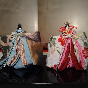 以下が含まれることがあります： カラフルな着物姿の伝統的な日本の女の子人形2体。人形は布製で、金と銀の装飾が施されています。人形は黒い台の上に座っています。