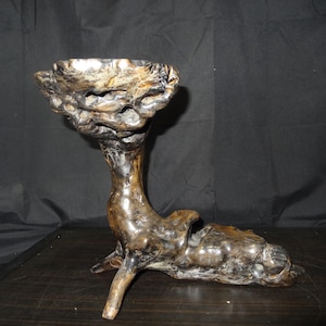 May include: A unique, handcrafted wooden sculpture with a cup-like top and a textured, organic design. The sculpture is primarily brown with lighter, natural wood tones, and stands on a dark surface.