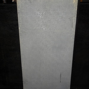 May include: A rectangular, light gray panel with a repeating, swirling pattern. The panel has a black border and appears to be part of a larger structure. The surface shows signs of wear and tear, with some discoloration and scratches.