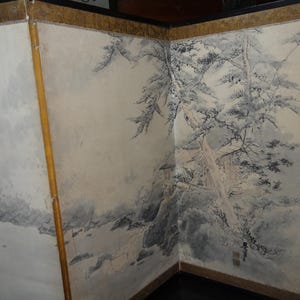 May include: A three-panel folding screen with a traditional Japanese landscape painting. The artwork features a monochrome depiction of trees, mountains, and a building, with gold trim and a dark wooden frame.