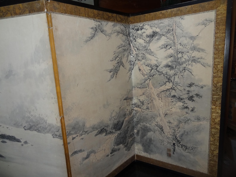 May include: A three-panel folding screen with a traditional Japanese landscape painting. The artwork features a monochrome depiction of trees, mountains, and a building, with gold trim and a dark wooden frame.
