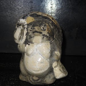 May include: A ceramic Tanuki statue, a traditional Japanese raccoon dog figure. The statue is primarily gray and cream-colored, with visible cracks and wear. It holds a sake bottle and has a friendly expression.