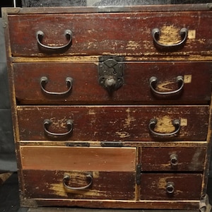 Könnte beinhalten: Eine rechteckige Holzkommode mit dunkelrotbrauner Oberfläche und sichtbaren Gebrauchsspuren. Sie verfügt über drei durchgehende Schubladen mit dunklen Metallgriffen, eine kleinere Schublade sowie zwei übereinanderliegende Seitenschubladen mit Knöpfen.