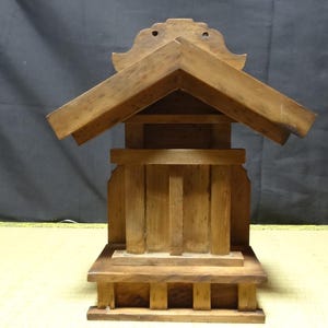 Puede incluir: Un pequeño modelo de santuario sintoísta de madera de color marrón claro que presenta un techo a dos aguas con una pieza decorativa en la cumbrera, una estructura central rectangular con listones verticales y una base escalonada de madera natural.