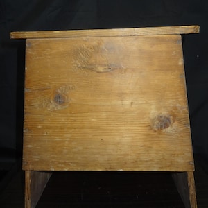 May include: A small, rustic wooden stool with a rectangular top and angled legs. The stool is constructed from light-colored wood, showcasing the natural grain and knots. The design is simple and functional, with a slightly overhanging top.
