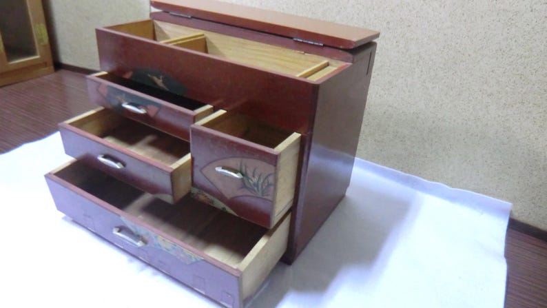 May include: A wooden jewelry box with a red finish and intricate floral designs. The box has four drawers and a hinged lid.