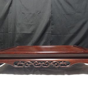 May include: A rectangular dark brown wooden display stand with a smooth flat top surface and a raised border. The stand features four curved legs connected by an intricate openwork apron with carved scroll patterns along the front side.
