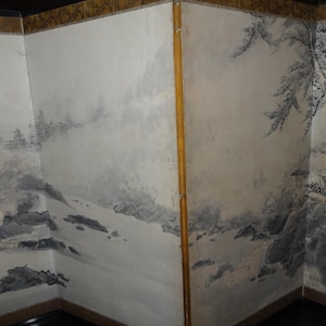 May include: Corner of a room with painted walls depicting a landscape scene in shades of gray and black. The artwork features mountains, trees, and a small building, creating a serene, artistic ambiance. The walls are bordered with a gold trim.