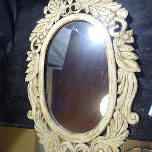 以下が含まれることがあります： クリーム色の装飾的なフレームが付いた楕円形の鏡。 フレームには、彫刻された花と葉のデザインが施されています。 鏡の表面は周囲の環境を反射します。 全体的なスタイルはヴィンテージまたはアンティークです。