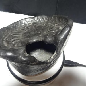 May include: A dark charcoal grey ceramic oil warmer featuring a wide, shallow bowl with a textured, scalloped rim and a central circular indentation. The base of the bowl has a large, arched opening for heat access, and a black power cord is attached.
