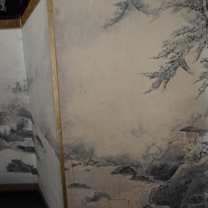 May include: A section of a traditional Japanese screen featuring a landscape painting. The artwork depicts a serene scene with mountains, water, and trees in shades of gray and black against a beige background. The screen has a gold border.