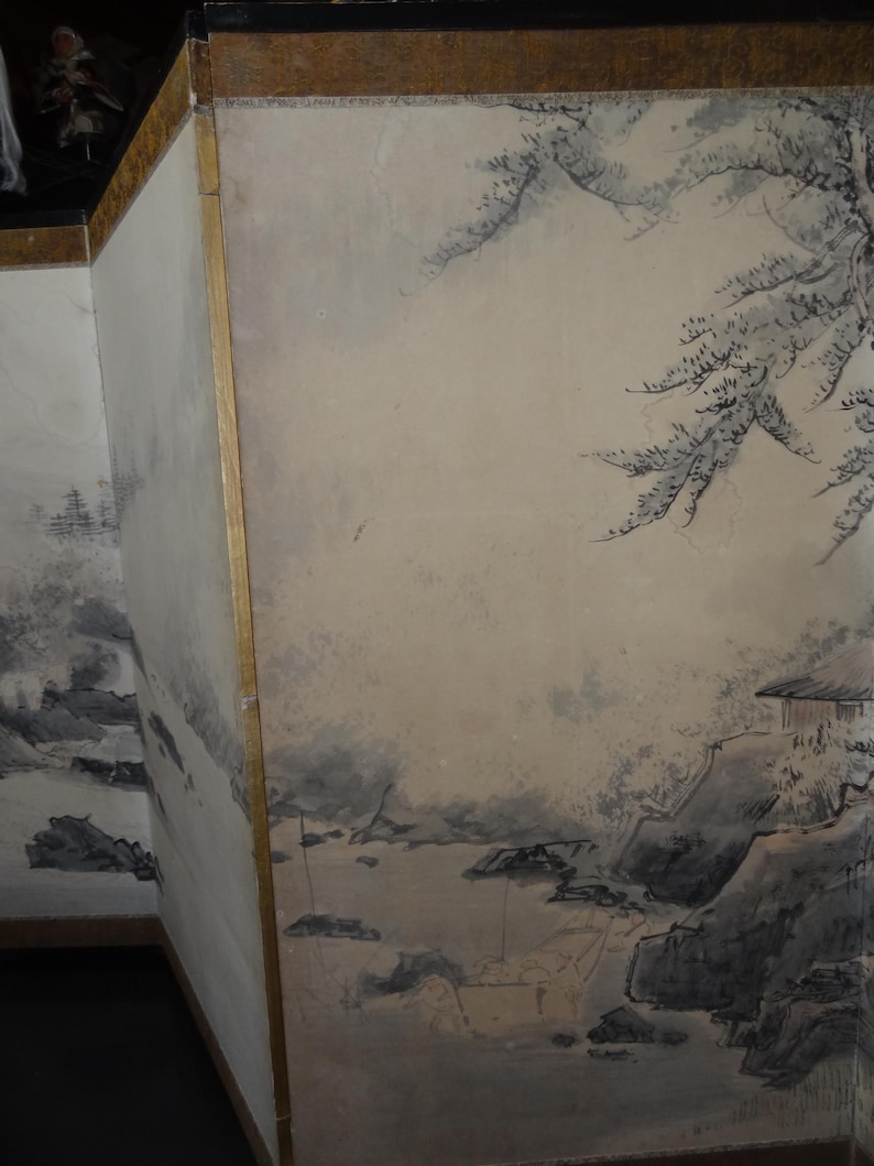 May include: A section of a traditional Japanese screen featuring a landscape painting. The artwork depicts a serene scene with mountains, water, and trees in shades of gray and black against a beige background. The screen has a gold border.