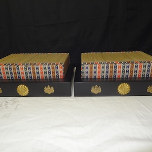 Peut inclure: Deux boîtes rectangulaires en bois noir avec des accents floraux dorés. Chaque boîte a un dessus à motifs avec des motifs géométriques dorés, rouges, bleus et blancs. Les boîtes sont exposées sur une surface blanche sur fond noir.