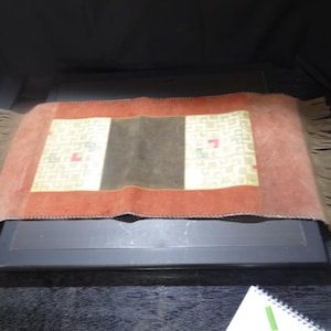 May include: A rectangular, decorative rug with a central brown panel, bordered by a rust-colored frame. The rug features a cream-colored section with small, colorful geometric patterns. The edges have a fringe.