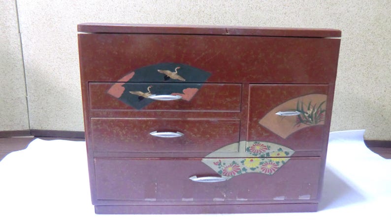 May include: A red wooden chest with four drawers. The drawers have silver handles and are decorated with Japanese-inspired designs, including cranes, fans, and flowers.