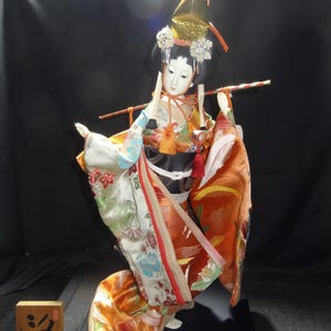 Puede incluir: Muñeca japonesa con kimono tradicional, tocado dorado y sosteniendo un palo largo. El kimono presenta estampados florales en naranja, blanco y azul. Un letrero de madera con caracteres japoneses está frente a la muñeca.