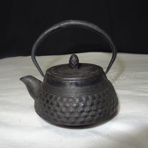 Puede incluir: Una pequeña tetera de hierro fundido negro con un asa curva y un pico. La tapa está rematada con un pomo decorativo. El cuerpo de la tetera tiene un diseño texturizado y estampado. La tetera está sobre una superficie blanca.