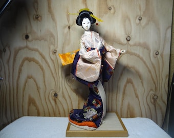 May include: A traditional Japanese doll wearing a colorful kimono with intricate patterns. The doll has a white porcelain face with black hair and is posed in a dancing position.