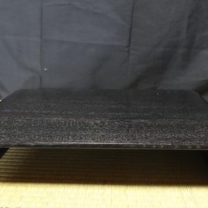 May include: A rectangular black display stand with a smooth, dark finish and curved, scroll-like ends. The stand features a flat top surface and a low-profile design, constructed with a solid, continuous structure that curls inward at both sides.
