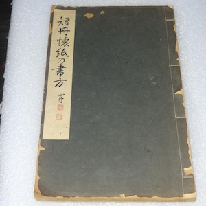 以下が含まれることがあります： 濃い灰色の表紙と、日本語のテキストが書かれたクリーム色のラベルが付いた、ヴィンテージの日本の本。本の端は摩耗と経年劣化の兆候を示し、色あせた外観です。ラベルには縦書きのテキストと赤い印章があります。