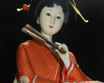 May include: A Japanese doll with a serene expression, wearing a vibrant orange kimono with white and gold wave patterns. The doll has black hair, a decorative hairpiece, and holds a small fan. The background is black.